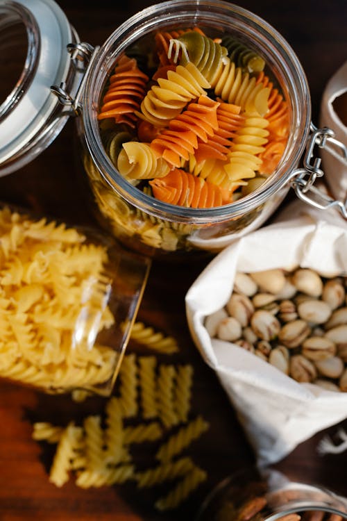 Top view of glass jars with uncooked fusilli and rotini pasta placed on table near ECO friendly bag with pistachios