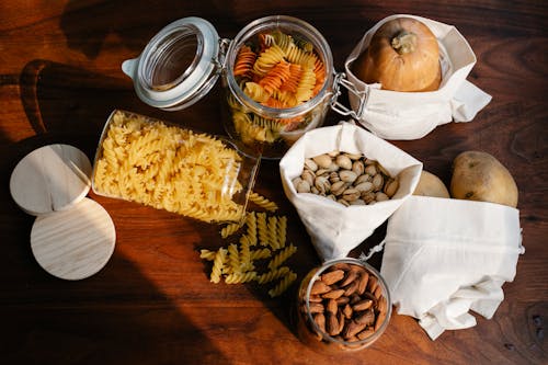 Top view jars of raw pasta placed on wooden table near ECO friendly sacks with pistachios and almonds near pumpkin and potatoes