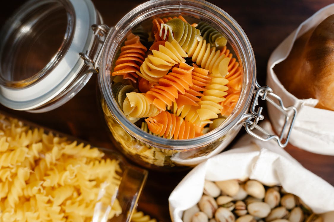 From above glass jar with colorful rotini placed on table near fusilli in container and pistachios and bread in bags
