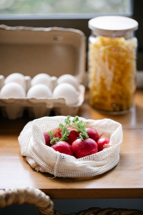 Organic fresh radishes in reusable bag placed on kitchen table near box with eggs