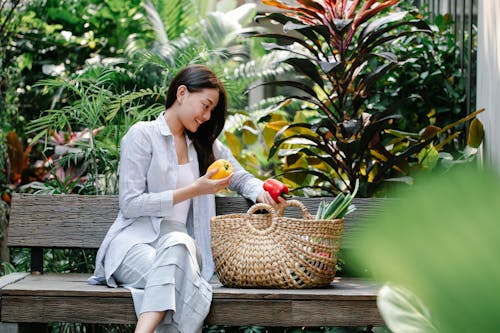 Content Asian female wearing casual outfit placing fresh yummy capsicums in wicker basket while sitting on wooden bench in verdant garden