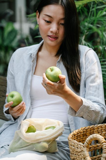 Crop young Asian female collecting fresh green apples in reusable bag while sitting in verdant garden during harvest season
