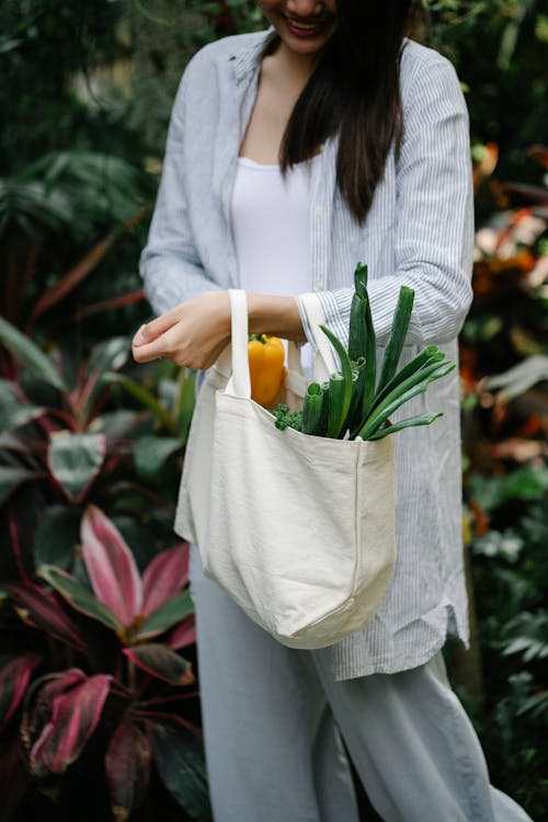 Crop happy female wearing casual clothes carrying textile bag with ripe fresh vegetables in lush summer garden