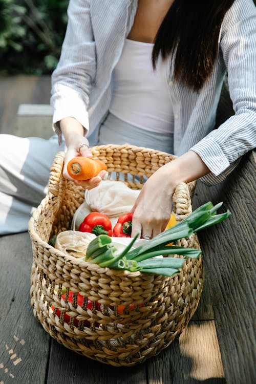 Crop anonymous female wearing casual outfit placing fresh organic vegetables in wicker basket while sitting on wooden bench in lush garden