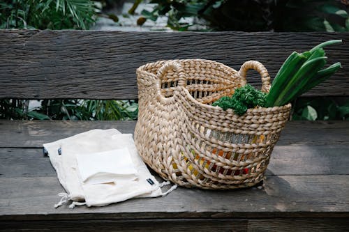 Free Wicker basket with heaped organic vegetables placed on wooden bench near reusable grocery bags in lush garden Stock Photo