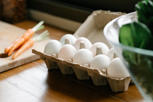 Carton box with white organic eggs placed on kitchen counter near chopping board with carrots