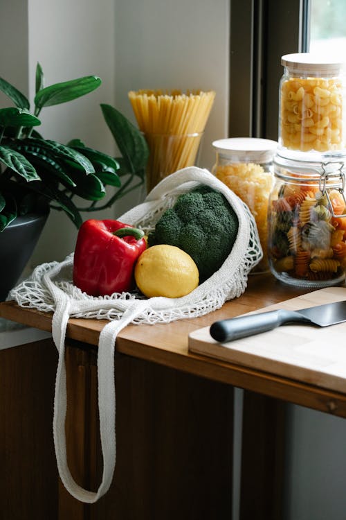 Composition of ripe fresh vegetables in net bag placed on kitchen counter near jars with pasta and chopping board with knife