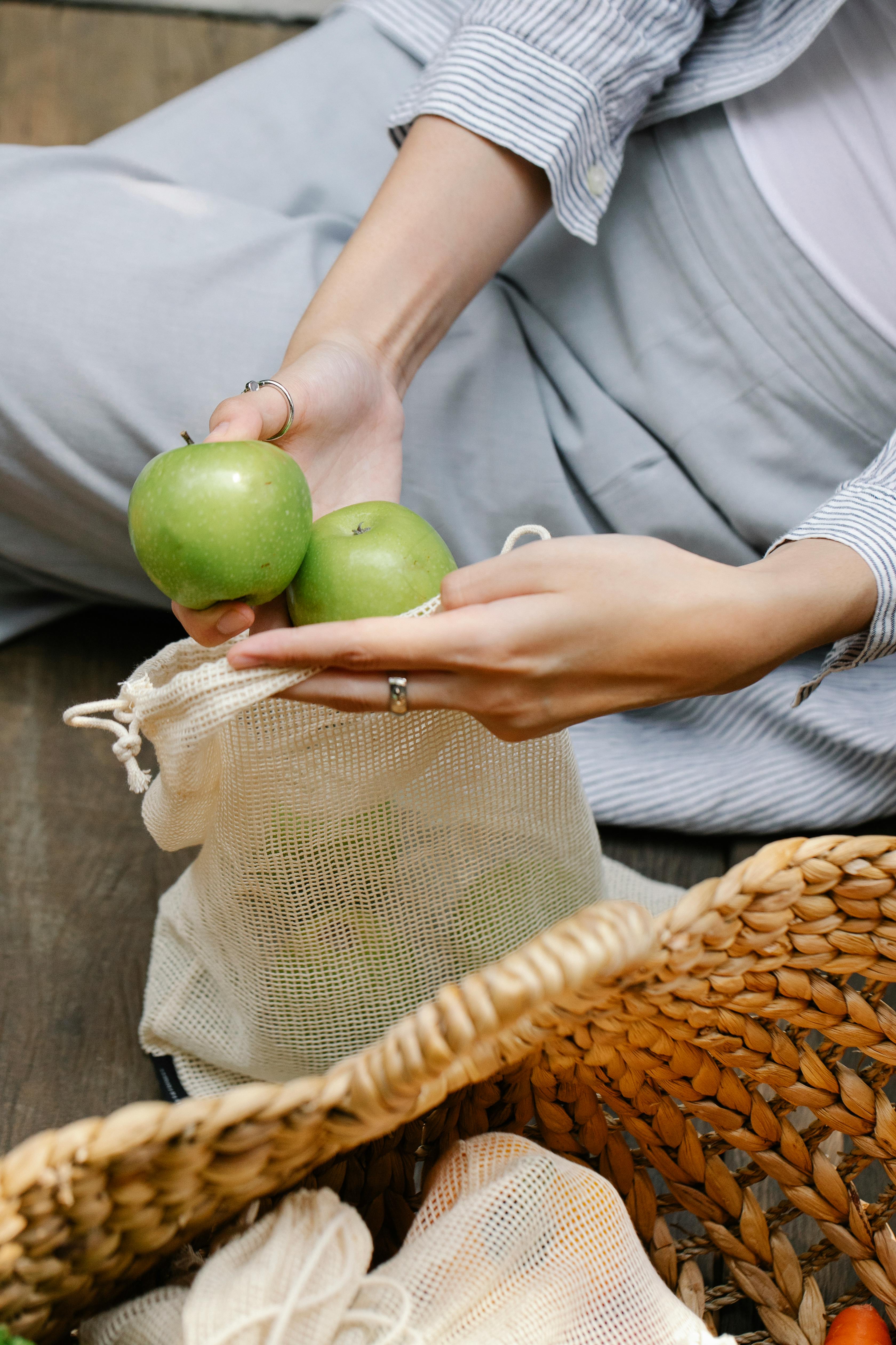 crop faceless woman placing green apples in reusable bag