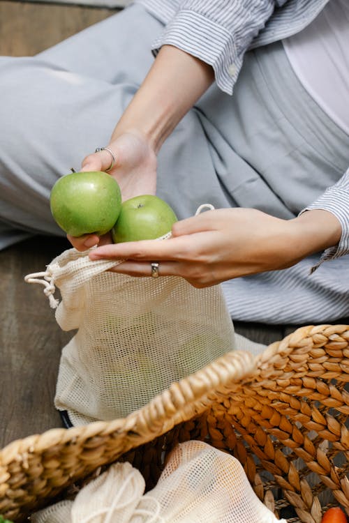 Crop faceless woman placing green apples in reusable bag