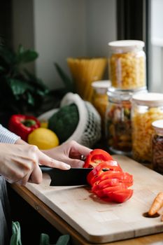 Crop unrecognizable female cutting red bell pepper on wooden chopping board while preparing healthy salad in kitchen