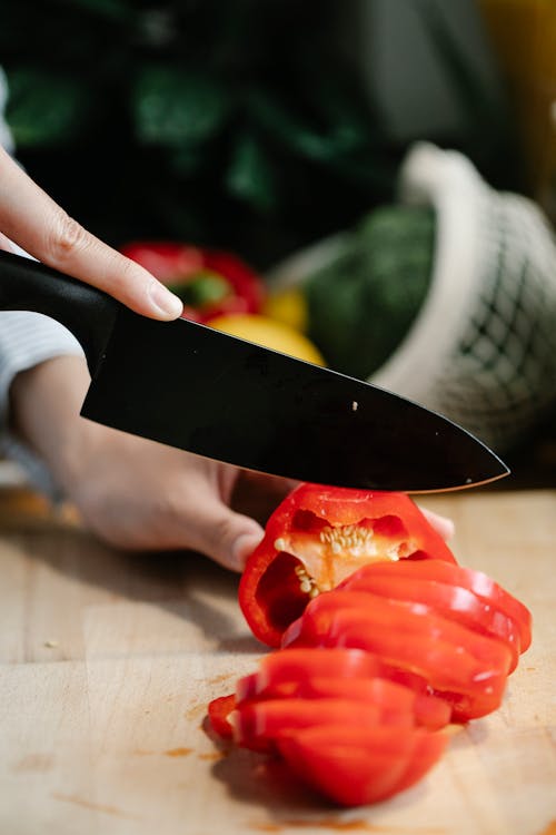 Crop anonymous woman chopping healthy red bell pepper with knife on cutting board while cooking in kitchen at home