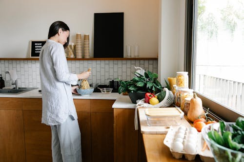 Side view of young Asian female with long dark hair in comfy clothes preparing noodles while standing at counter with assorted fresh products in kitchen