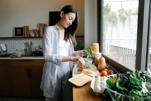 Side view of Asian female adding fresh parsley into glass container with pasta and bolognese sauce while cooking in kitchen with various products and kitchenware