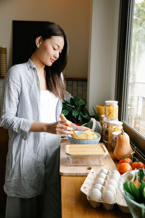 Free Side view of content Asian female standing at counter with bowl of spaghetti and various products while cooking in kitchen Stock Photo