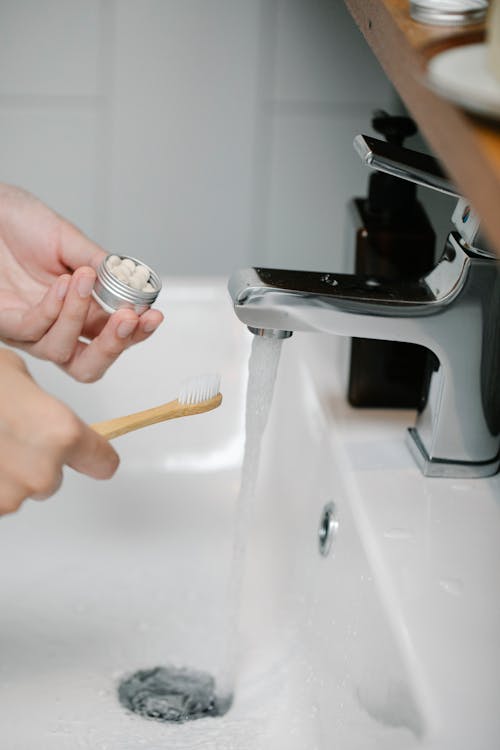 Crop anonymous person with toothpaste tablets standing near sink with pouring water while brushing teeth in bathroom during daily hygiene routine