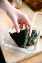 Crop anonymous cook putting onigiri wrapped in nori into glass container placed on cutting board while cooking at counter in kitchen