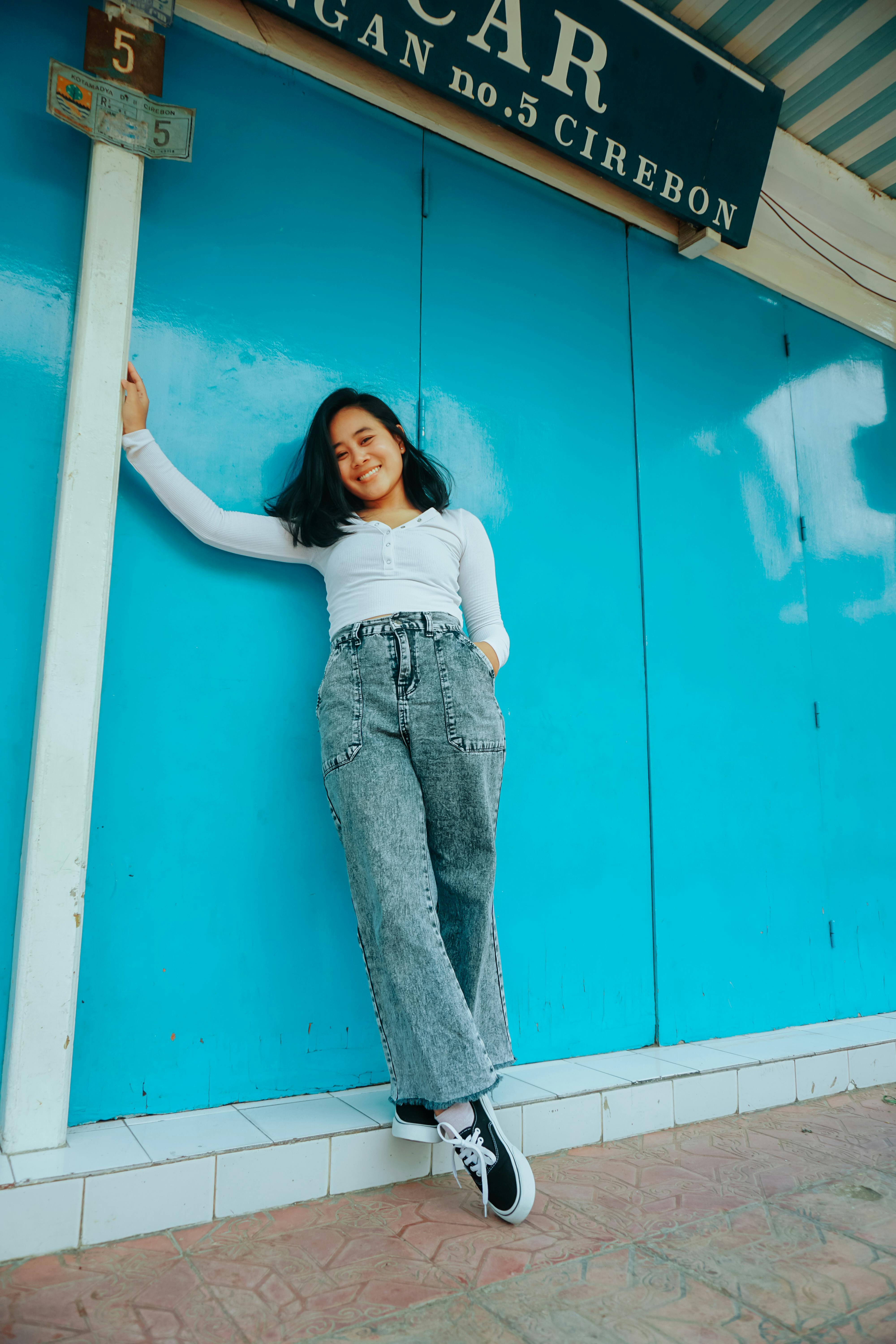 Full body of smiling Asian female in casual clothes dangling foot from tiled edge while standing near blue wall outside and looking at camera