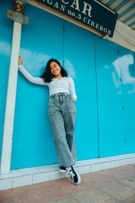 Full body of smiling Asian female in casual clothes dangling foot from tiled edge while standing near blue wall outside and looking at camera