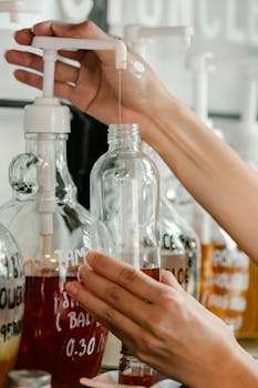 Crop anonymous person pouring colorful soap from big dispenser on shelving into reusable bottle