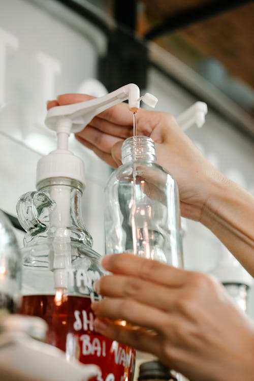 Crop unrecognizable person pouring red liquid soap into transparent reusable bottle from dispenser