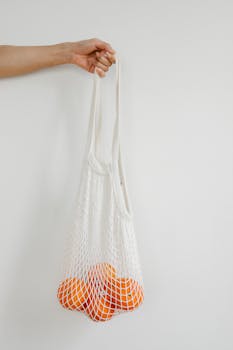 Hand holding a reusable mesh bag filled with fresh, ripe oranges against a white background.