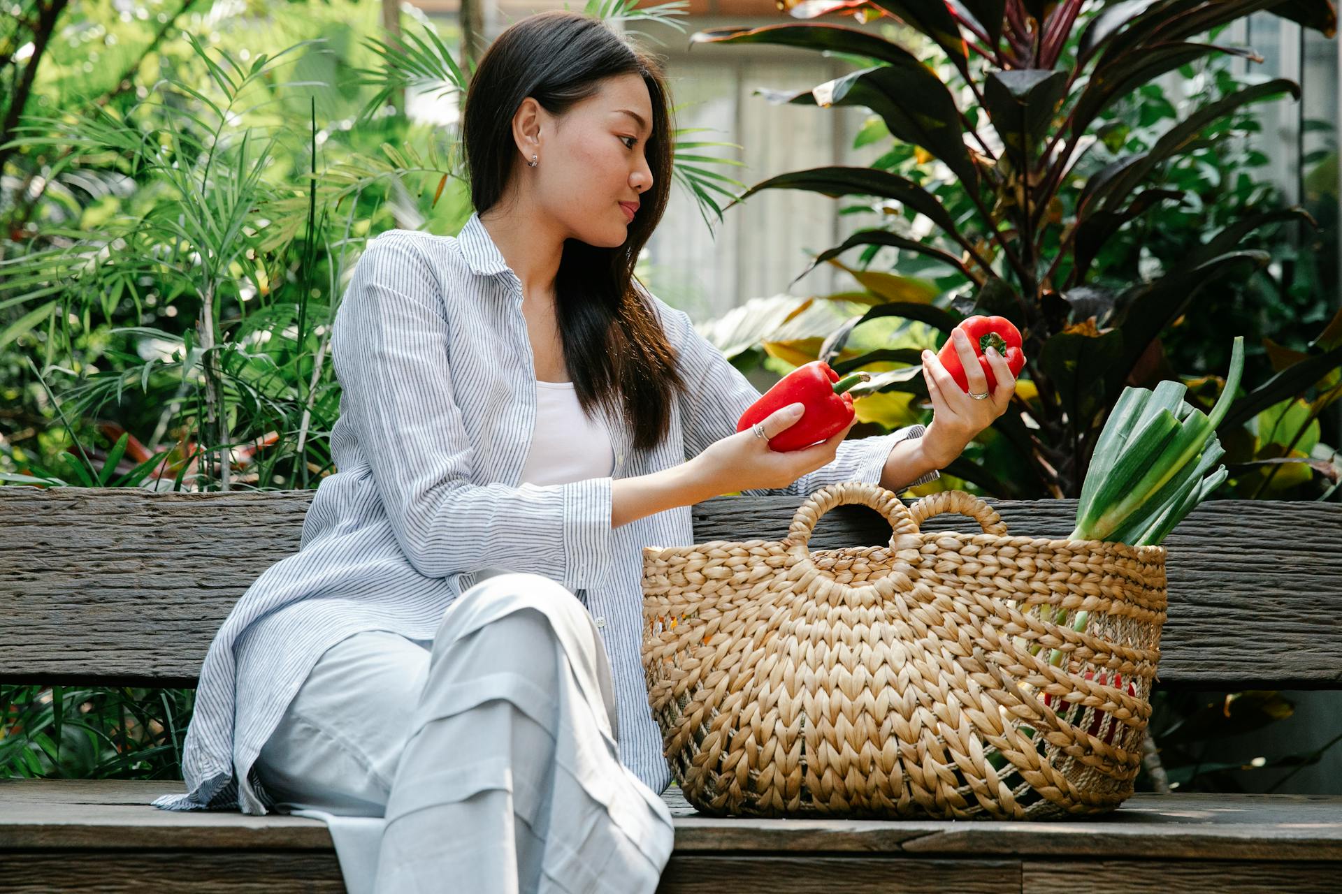 Calm Asian female in casual clothes sitting on wooden bench among green plants and putting ripe red peppers in wicker basket in daytime