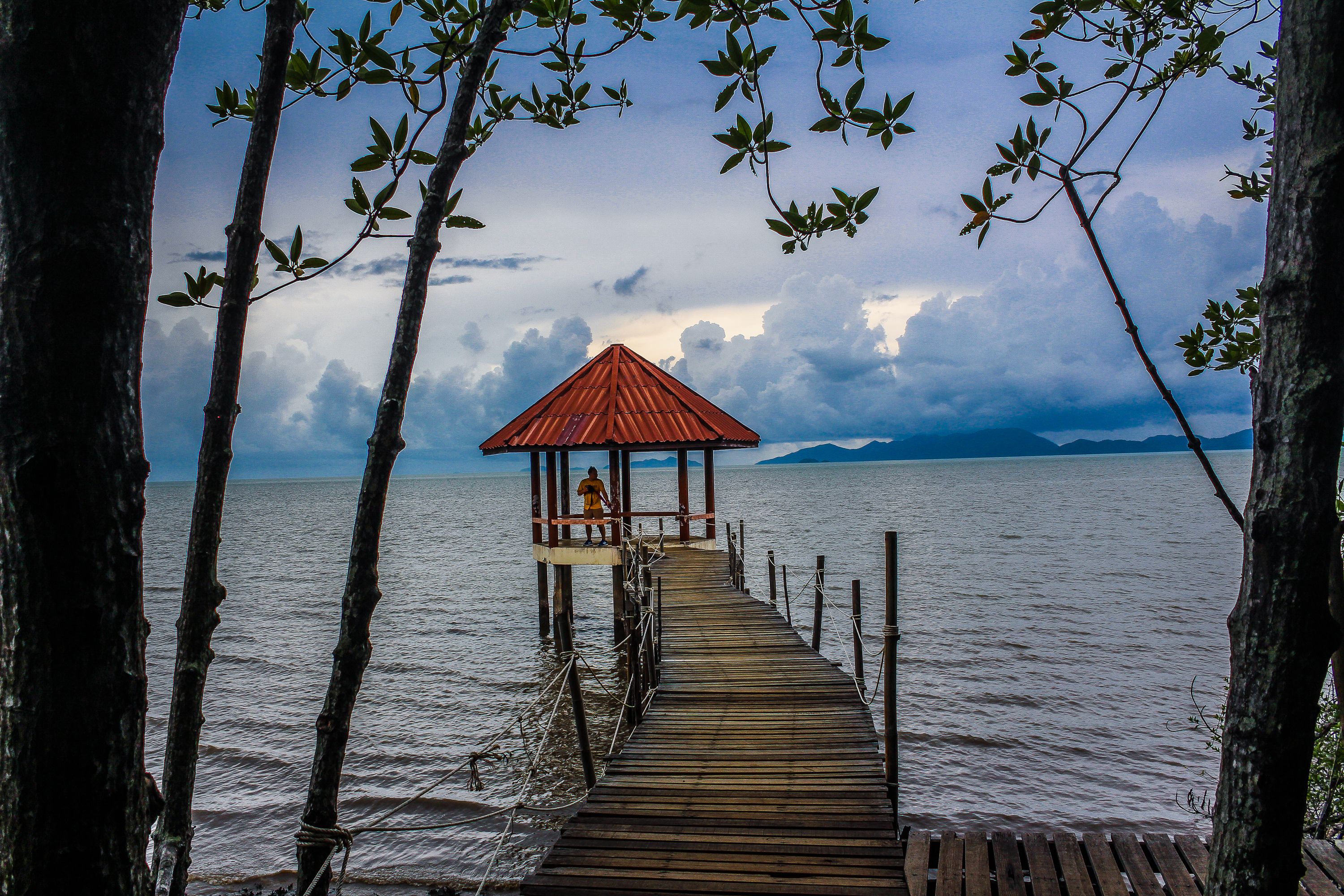 Peaceful boardwalk leading to a gazebo overlooking the ocean at sunset, framed by trees and dynamic clouds.