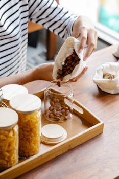 Crop female putting fresh almonds from bag to glass jar placed on wooden tray with jars with macarons in light room