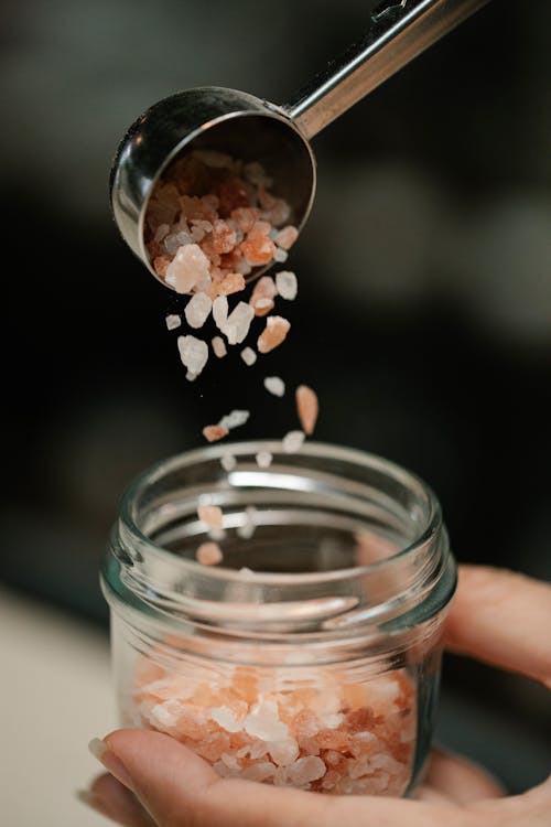 Free Crop anonymous female pitting aromatic healthy salt for bath in small glass jar against blurred background Stock Photo