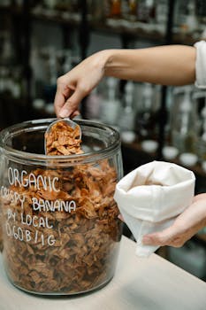 Hand scooping organic crispy banana chips from a jar indoors.
