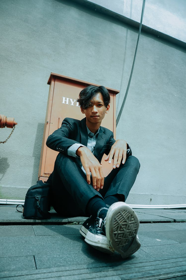 Ethnic Teenager In Gray Suit Sitting Near Concrete Wall