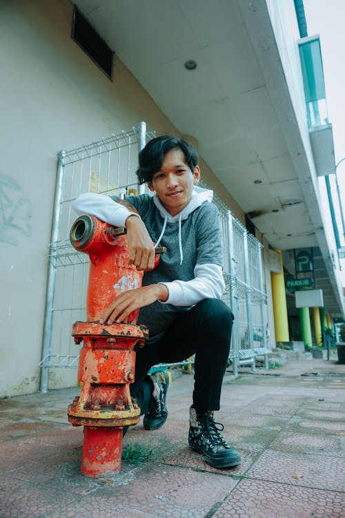 Free Full body of positive Asian male teenager in trendy outfit sitting on haunches ad leaning on shabby red fire hydrant on city street Stock Photo