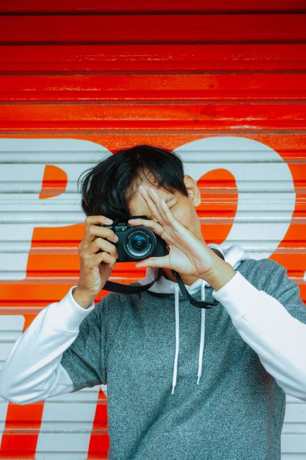 Unrecognizable young Asian male photographer in stylish outfit taking pictures on modern camera while standing near red metal rolling door with white inscription