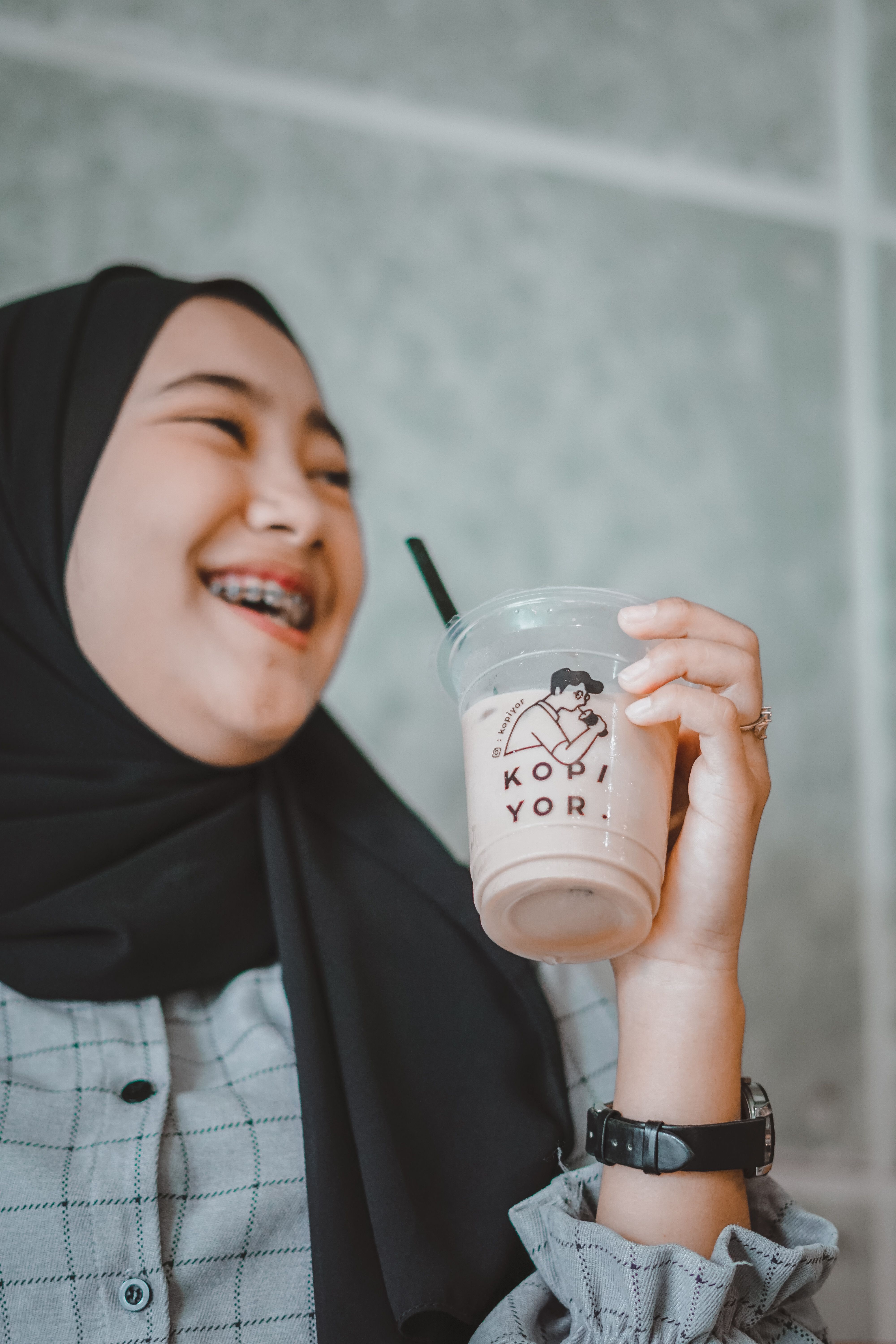 Crop cheerful Muslim female teenager in casual clothes and hijab drinking cold takeaway beverage and laughing happily in cafeteria