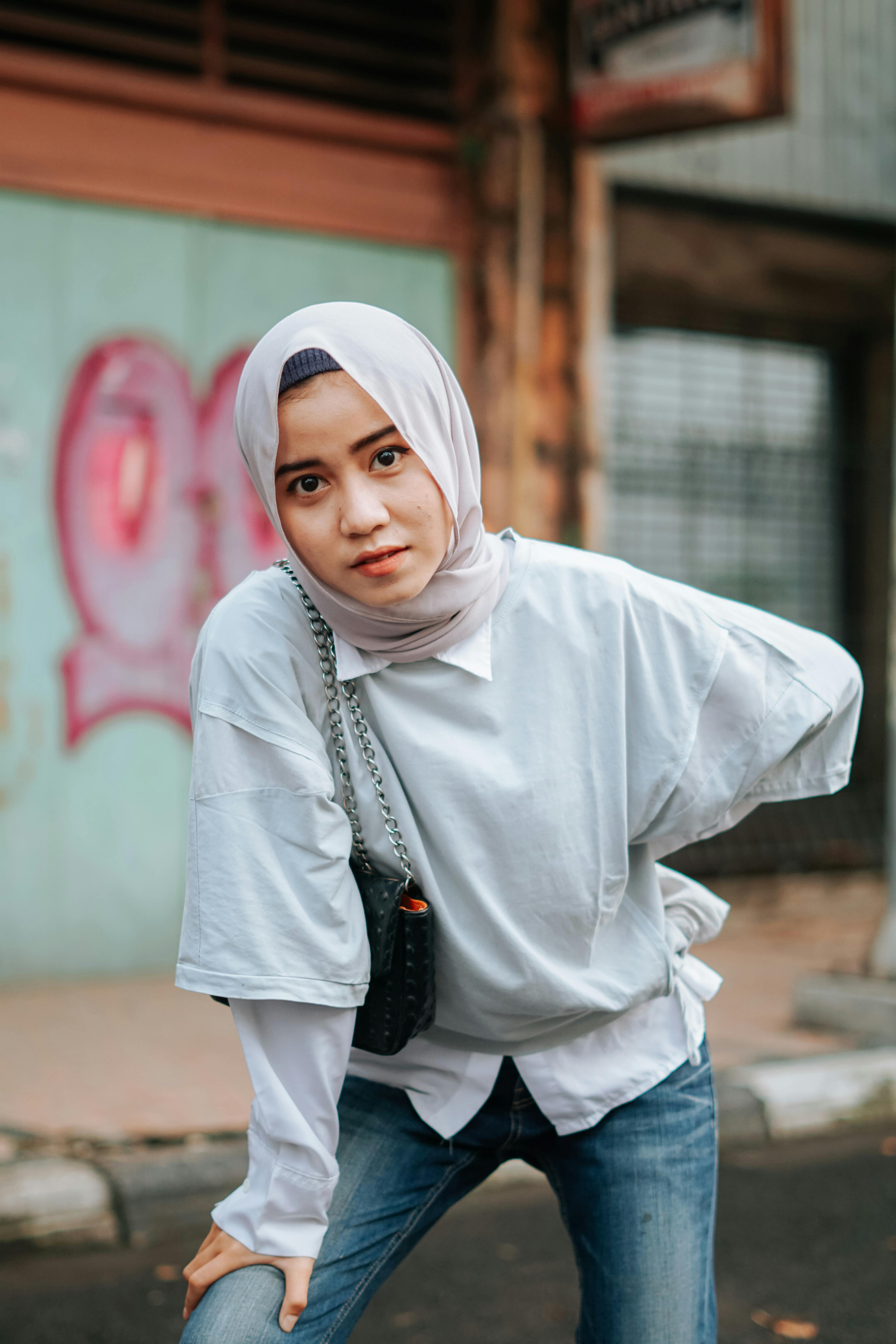 Serious young Asian lady in casual clothes and traditional Muslim hijab leaning forward and looking at camera while standing on street in daytime