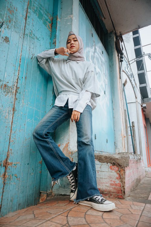 From below of young slender Muslim female in headscarf looking at camera near shabby building