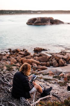 Man controls drone using tablet while sitting on rocky coastline overlooking the sea.