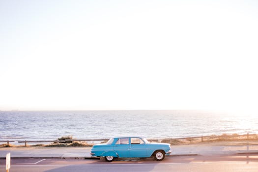 A classic blue vintage car is parked by the seaside with a scenic ocean view during sunset.