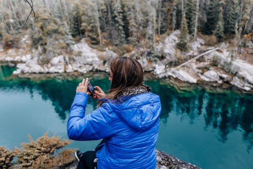 Woman in Blue Jacket Taking Photo of Lake