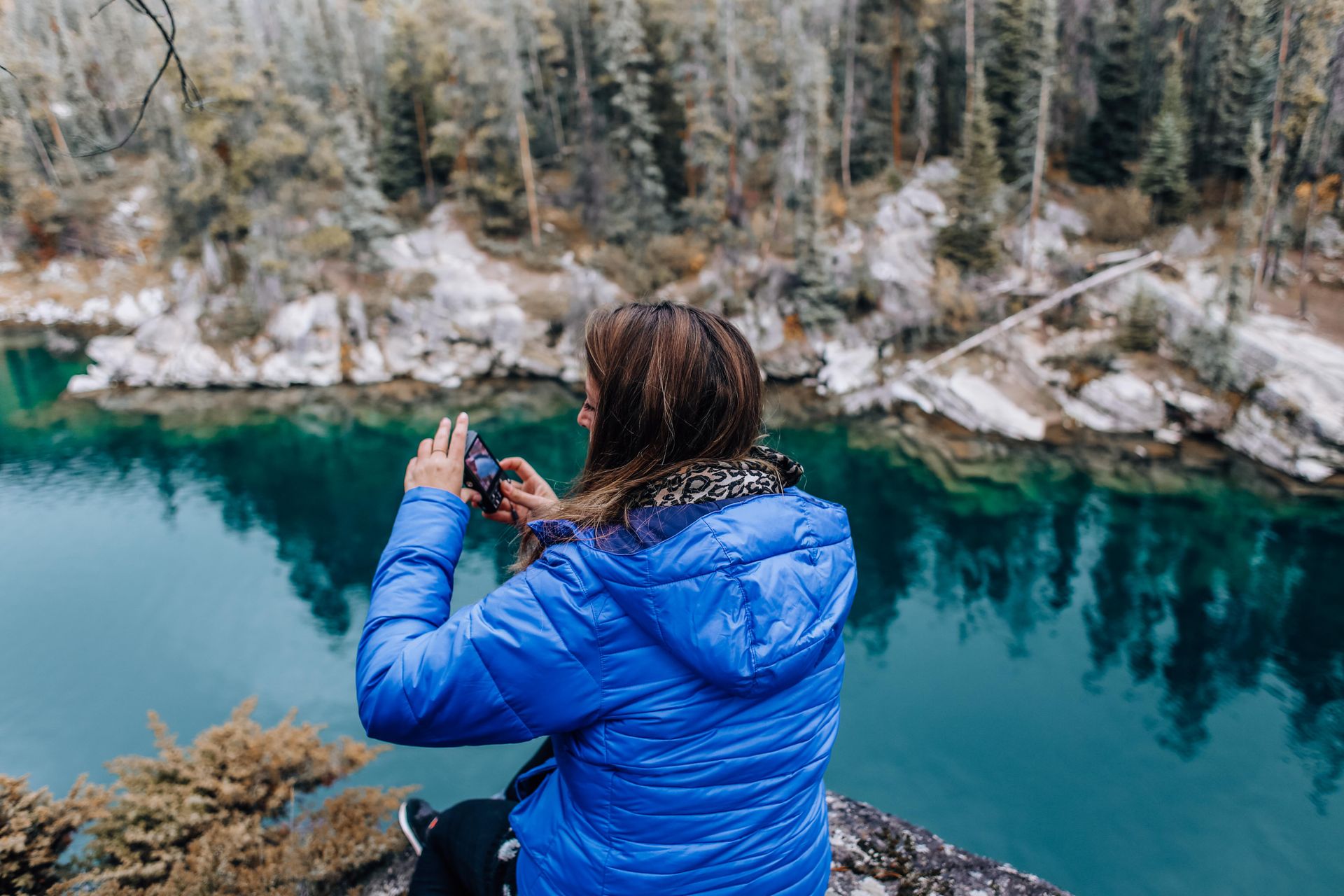 A woman in a blue jacket captures the beauty of a lake and forest on her camera.