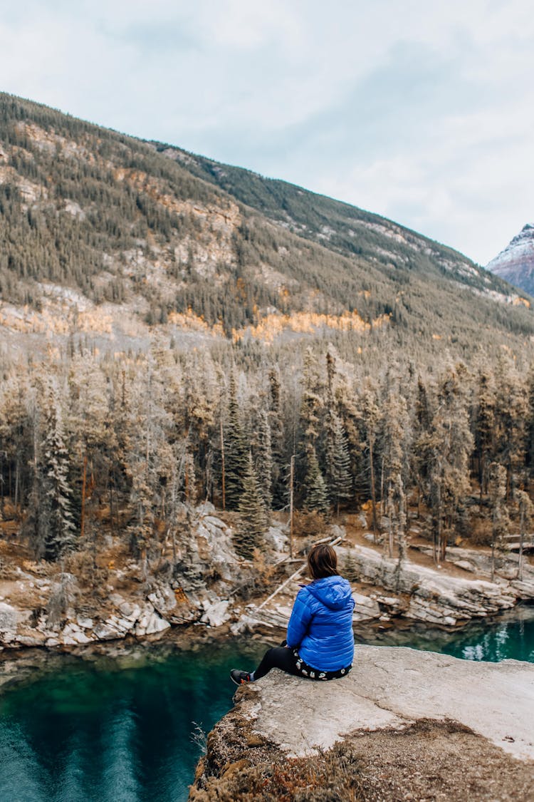 Person In Blue Jacket Sitting On Rock Cliff 