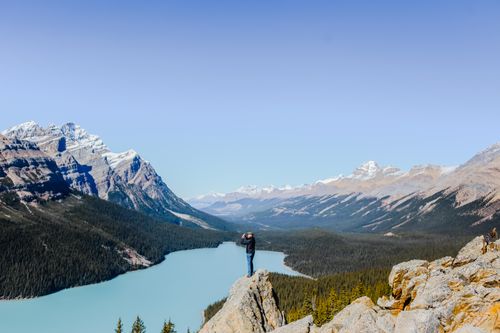 Free A lone hiker stands on a rock overlooking a stunning mountain lake and forest landscape. Stock Photo