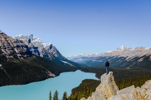 Free A person stands on a cliff, enjoying a scenic view of a mountain lake and peaks. Stock Photo