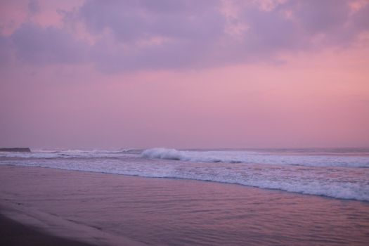 Serene beach scene with pink sky at sunset, waves gently crashing on shore.