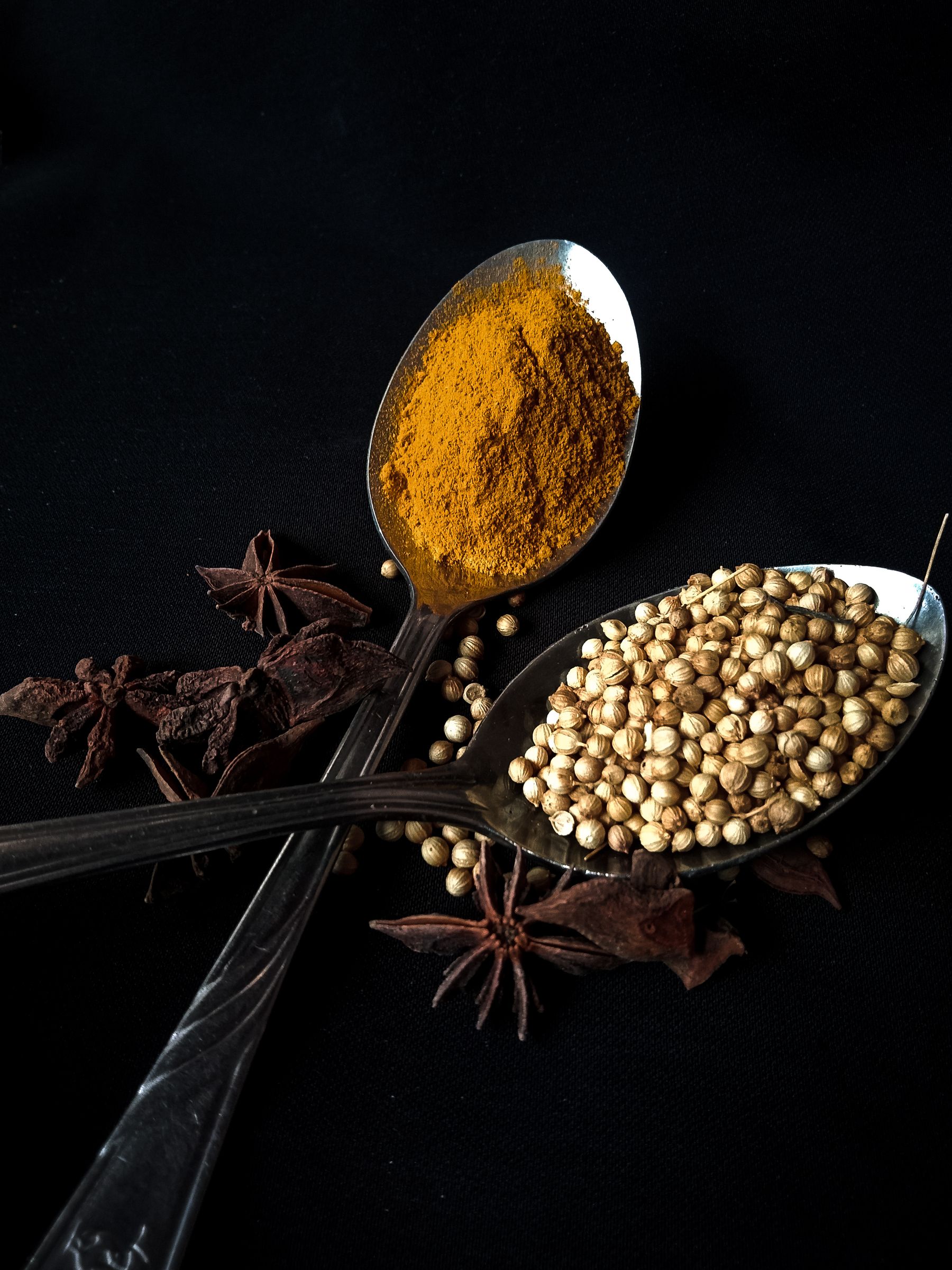 Turmeric, coriander and star anise arranged on dark spoons against a black backdrop