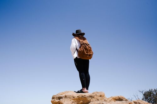 Free A woman in a hat stands on a cliff edge with clear blue skies, wearing a backpack. Stock Photo