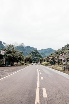 A picturesque asphalt road with lush green mountains and rural houses in the background.