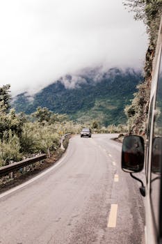 A vehicle navigates a winding road through a misty mountainous countryside, surrounded by lush greenery.