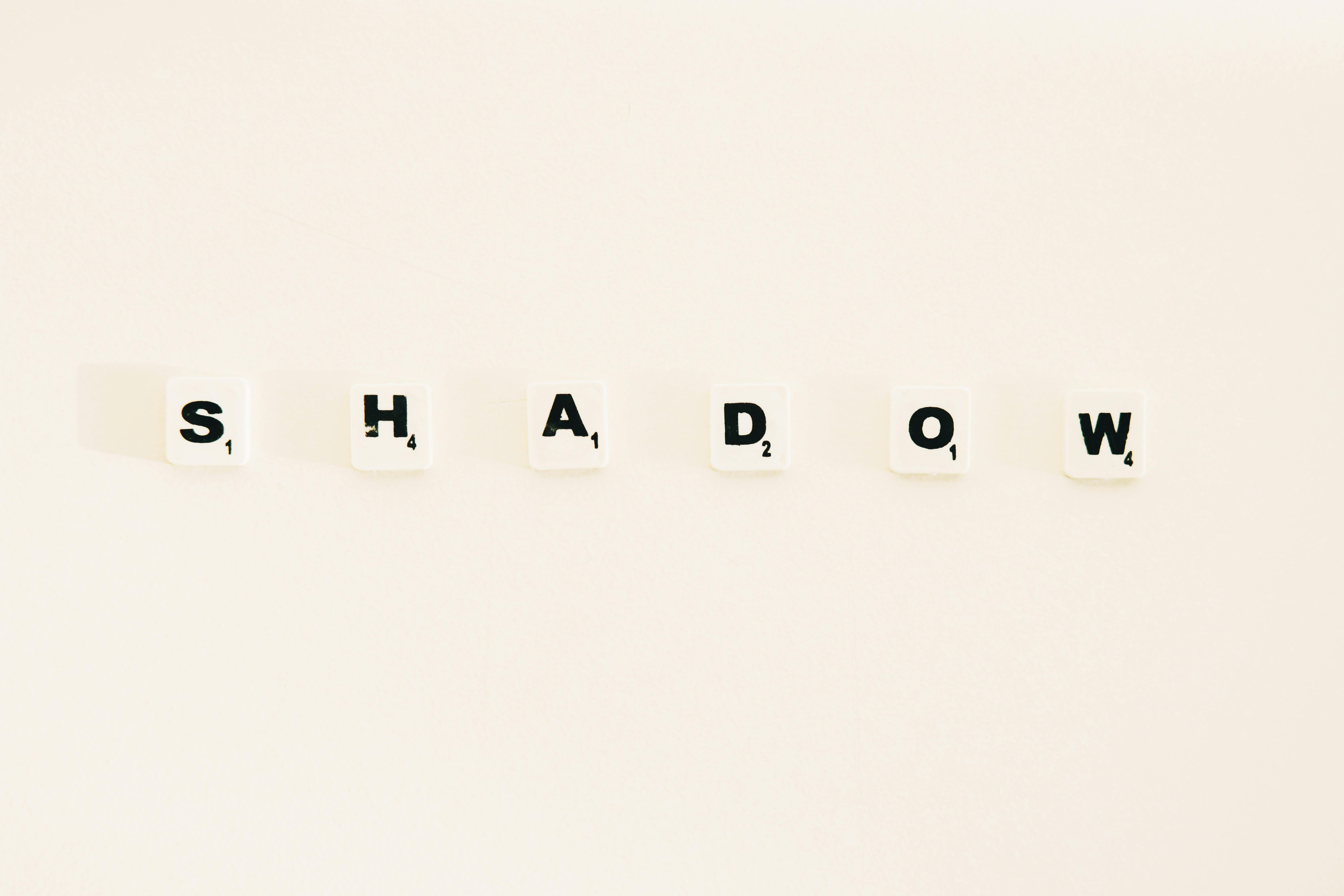 Scrabble tiles spelling 'SHADOW' on a white background with light shadows.