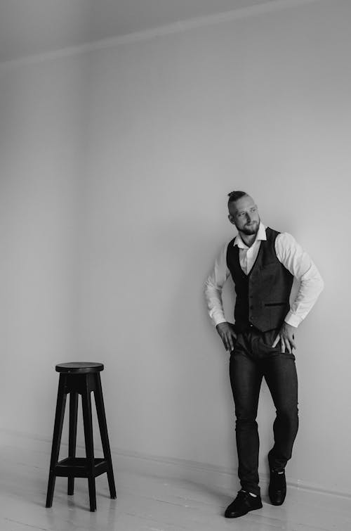 Man in White Long Sleeve Shirt and Black Vest Standing Beside Black Wooden Chair
